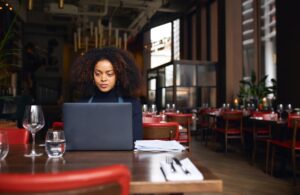 A woman with curly hair sits at a wooden restaurant table while focused on her laptop. The background shows an elegantly designed dining area with red chairs, set tables.