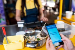 A person holds a smartphone displaying a successful "Online Banking" payment confirmation in front of a street food stall. In the background, a vendor in a blue apron stands behind a counter featuring pots of skewered food and a payment terminal.