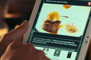 A person’s hand holds a tablet displaying a menu item for Bananas Foster Bread Pudding, complete with a photo of the dessert topped with gelato. The screen includes a descriptive blurb about the dish and a section for recommended wine pairings.