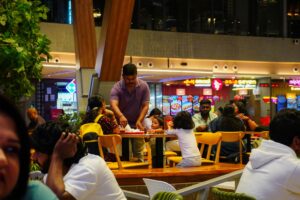 Families and diners gather at a busy food court, with various restaurant signs like "Asia Seven" visible in the background. In the center, a man serves a meal to children at a table, highlighting a casual and lively dining atmosphere.