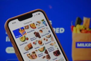 A smartphone displays the "Categories" screen of the Milkrun grocery delivery app, featuring various food sections like "Fruit & Veg" and "Meat & Seafood." The phone is held against a bright blue background that includes a blurred image of a grocery bag.