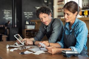 Two restaurant workers in aprons are sitting at a counter, carefully reviewing receipts and financial documents. They are using a tablet and a calculator to manage their bookkeeping in a rustic, dimly lit establishment.