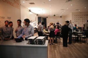 Staff members in checkered shirts gather at a host stand in a busy, modern restaurant. Customers are seated at various tables in the background under bright, warm lighting.