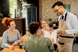 A smiling waiter in an apron presents an open menu to a group of three friends seated at a wooden cafe table. The customers, including two women and a man, appear engaged and cheerful as they prepare to order.