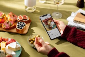 A person in a maroon sweater holds a smartphone displaying a social media post of a cheese and fruit plate while reaching for an olive from a small bowl. The table is spread with a variety of snacks, including tapas, charcuterie, and wine, set against a warm, sunlit background.