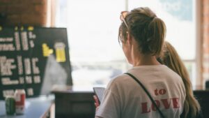 A woman with her hair in a ponytail stands in a cafe, looking toward a large chalkboard menu. She holds a smartphone while wearing a white t-shirt featuring bold red text on the back.