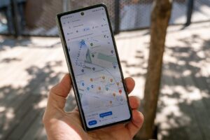 A person's hand holds a slim, modern smartphone displaying a Google Maps navigation route near Chelsea Market in New York City. The device is held outdoors on a sunny day, with the blurred background showing a wooden walkway and shadows from nearby trees.