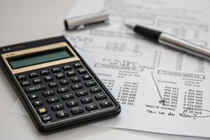 A black financial calculator sits on top of detailed investment papers covered in rows of numbers and handwritten notes. A silver pen with its cap removed rests nearby, suggesting an active session of budgeting or financial planning.