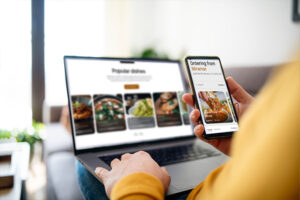 A person is simultaneously browsing a restaurant's "Popular dishes" on a laptop and using a smartphone to view an ordering interface for Miramar. The image highlights a seamless multi-device experience for selecting and ordering food online.