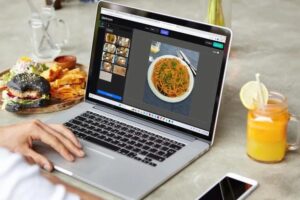 A person is using a laptop at a table to edit a photo of a pasta dish using an online software called Darkroom. Real food, including a burger and a glass of orange juice, sits on the table surrounding the computer.