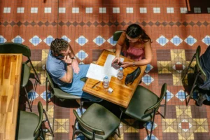 The second image shows a man and a woman sitting at a wooden table on a vibrantly patterned tile floor, both preoccupied with their phones.