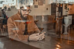 A man with a beard and glasses sits at a wooden table in a cafe, focused on his smartphone. The shot is captured through a window, creating soft reflections of the street and interior decor over the scene.