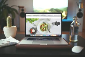 A MacBook Pro sits open on a desk, displaying a website featuring a healthy salad bowl and the German text. The desk is arranged with a small potted cactus, a glass bottle, and a newspaper, set against a softly blurred background of a living room.