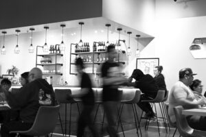 This black-and-white photograph captures a lively bar area where patrons sit at the counter and tables under a row of elegant pendant lights.