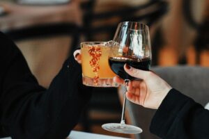 Two people are shown in a close-up shot, clinking a glass of red wine and a spiced orange cocktail together in a toast. The warm, blurred background suggests an intimate and cozy setting, likely a restaurant or lounge.
