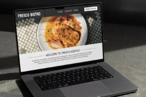 An open laptop displays the "Fresco Bistro" website, featuring a large image of a shrimp dish and a welcoming message about their Spanish cuisine.
