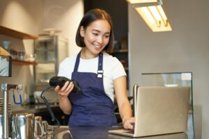 A smiling barista with a bob haircut stands behind a cafe counter while holding a card payment terminal. She is dressed in a white t-shirt and a navy blue apron, looking down at an open laptop on the counter.