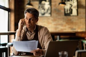A man wearing an apron sits at a table in a bar or restaurant while talking on his mobile phone. He appears focused as he reviews a stack of documents in front of his laptop.