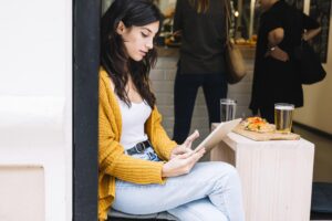 A woman in a yellow cardigan sits in a cafe while focused on a tablet in her hands. On the wooden table beside her, there is a glass of beer and a small plate of food.