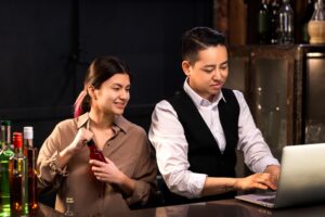 Two people work behind a bar, with one person typing on a laptop while the other holds a red glass bottle. Various bottles of liquor are lined up in the foreground against a dark, moody background.