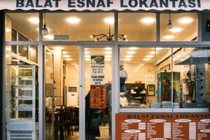 This image captures the storefront of "Balat Esnaf Lokantası," a traditional Turkish restaurant with a brightly lit interior and various menus displayed on the windows. Inside, a worker in a red apron stands near a vertical rotisserie, while a chef figurine and simple wooden tables are visible through the glass.