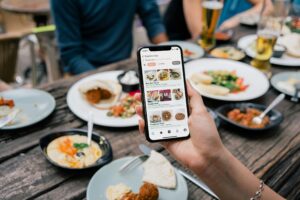 A person holds a smartphone displaying a food discovery app over a wooden table filled with various Mediterranean-style dishes and drinks. The background features a group of friends dining together at an outdoor restaurant setting.
