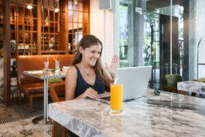 A woman sits at a marble table in a bright restaurant, waving at her laptop during a video call. She has a tall glass of orange juice in front of her, and the background features warm wooden shelving and large glass windows.