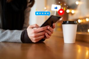 A person sits at a wooden table, holding a smartphone that displays colorful digital icons for ratings, likes, and social engagement. Next to them sits a white disposable coffee cup against a warm, blurred background of soft indoor lighting.