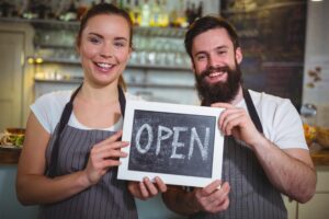 A smiling woman and a bearded man wearing aprons stand together in a cafe. They are both holding a small chalkboard sign that says "OPEN" in white lettering.