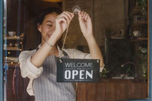 A young woman wearing a striped apron smiles as she hangs a "Welcome We Are Open" sign on the glass door of a shop. The warm lighting and wooden interior behind her suggest a cozy cafe or boutique atmosphere.