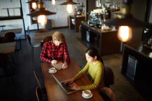 Two people work diligently at a long wooden table in a dimly lit, modern cafe. One person types on a laptop while the other writes in a notebook, both with warm drinks nearby.