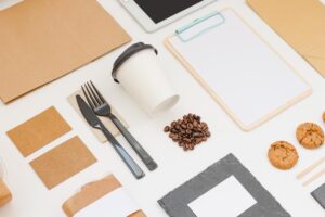 This high-angle flat lay features a collection of minimalist takeaway items, including a white coffee cup, black cutlery, and brown paper packaging arranged on a white surface. The composition is accented by scattered coffee beans, chocolate chip cookies, and a wooden clipboard with a blank sheet of paper.