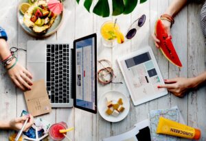 A vibrant flat-lay composition shows a wooden table scattered with travel essentials, including a laptop, tablet, maps, and tropical fruits. Hands are visible throughout the frame, engaged in activities like writing on a postcard, scrolling through a digital device, and enjoying a slice of watermelon.