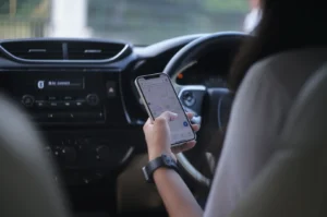 A person uses a smartphone to navigate GPS while driving, holding the phone near the steering wheel. The car's dashboard and controls are visible.
