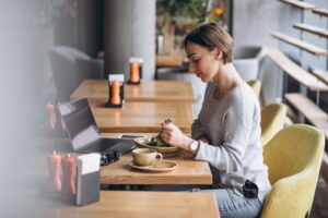 A woman with short brown hair sits at a wooden cafe table, focused on eating a meal while her open laptop and a cup of coffee rest nearby. The setting is bright and modern, featuring large windows and lime green chairs that create a clean, professional atmosphere.