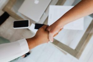 This high-angle shot captures two people shaking hands over a light-colored wooden table. The minimalist workspace includes a laptop, a smartphone, and documents, suggesting the successful conclusion of a business meeting or agreement.