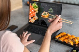 A woman uses a laptop to browse a sushi restaurant's website while holding a piece of maki with wooden chopsticks. Beside the computer, a plastic takeaway container is filled with a variety of brightly topped sushi rolls.