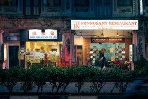 The image shows two illuminated restaurant storefronts, Shun Feng Kitchen and Ponnusamy Restaurant, nestled in a traditional building at night. A row of dark green bushes lines the sidewalk in the foreground as people pass by the open entrances.