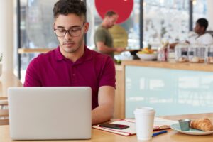 A young man wearing glasses and a maroon polo shirt focuses on his laptop while sitting at a wooden table in a bright cafe. Beside him, a coffee cup, a notebook, and a croissant on a plate complete his workspace while other patrons and a counter are visible in the blurred background.
