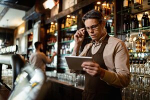 A male barista in a brown apron stands behind a bar, adjusting his glasses while focused on a digital tablet. In the background, a coworker is visible working near shelves stocked with various bottles and glassware.