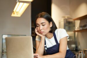 A woman wearing a dark blue apron looks intently at a laptop screen while resting her chin on her hand. She appears to be working in a cafe or shop setting with warm lighting overhead.