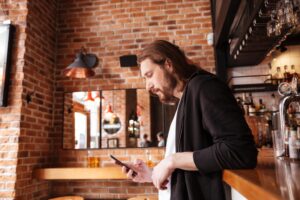 A man with long brown hair and a beard leans against a wooden bar counter while looking down at his smartphone. The setting is a rustic pub with exposed brick walls, warm industrial lighting, and shelves of bottled spirits in the background.