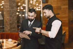 Two men in formal and smart-casual attire stand in a dimly lit, modern restaurant while discussing content on a tablet. The background features warm brick accents, soft bokeh lighting, and a circular wooden table set with a coffee cup.