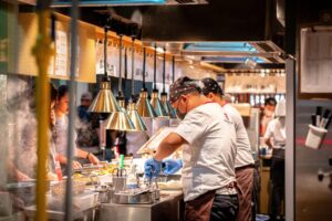 Inside a busy open-style kitchen, a chef wearing blue gloves and a patterned bandana carefully prepares a meal under bright heat lamps. Other staff members work alongside him in the bustling environment, while steam rises from the food prep area near the waiting customers.