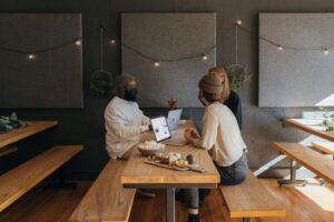 Three people wearing beanies and masks sit at a long wooden table in a modern cafe, working on laptops alongside a spread of snacks. The room features dark textured walls adorned with minimalist acoustic panels and a string of warm fairy lights.