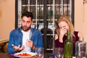 A young man gestures expressively while speaking as his female companion covers her face and laughs during an outdoor meal. A plate of pasta, wine bottles, and glasses sit on the wooden table before them against a backdrop of a decorative wrought-iron door.