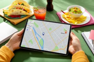 A person holds a tablet displaying a navigation map amidst a spread of Mexican food, including tacos, chips with guacamole, and a refreshing cocktail. The scene is set on a green tabletop, suggesting a moment of planning a route while enjoying a vibrant meal.