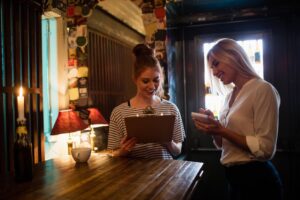 A young woman with her hair in a bun reviews a menu on a clipboard at a dimly lit restaurant table. Beside her, a waitress in a white shirt smiles while writing down the order on a small notepad.