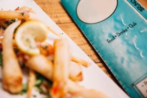 A close-up shot features a plate of crispy, golden spring rolls garnished with a fresh lemon slice and herbs. Next to the dish, a teal-colored menu with the words "Food + Drinks + Club" rests on a rustic wooden table.