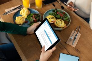 A person holds a tablet displaying an email inbox over a wooden cafe table set with brunch dishes like eggs Benedict and orange juice. Another person sits across from them, partially visible while using a smartphone.